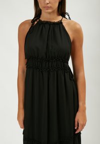 Vestido negro de cuello halter con cintura fruncida y acentos de volantes. Tejido suave con una silueta fluida. Se ata en los hombros.