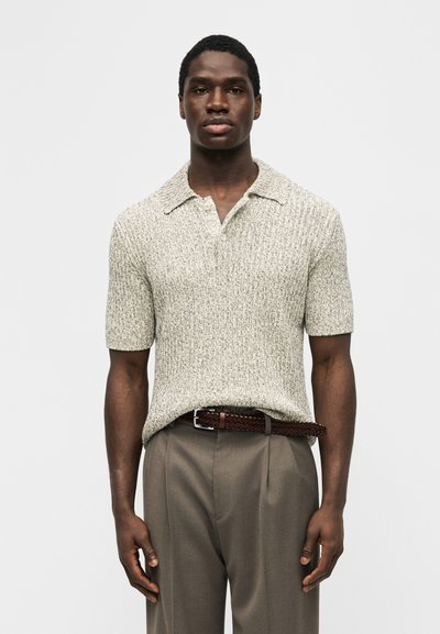 Άνδρας που φοράει μπλουζάκι πόλο με κοντά μανίκια και υφή μπεζ, κρυμμένο μέσα σε καφέ παντελόνια με ψηλή μέση και πλεκτή ζώνη, στέκεται μπροστά σε απλό φόντο.