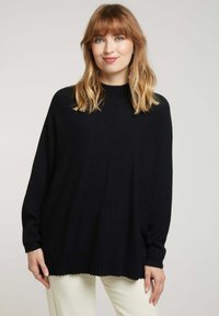 Pull noir oversize avec un col montant et des poignets côtelés. Texture douce et design à épaules tombantes, associé à un pantalon de couleur claire.