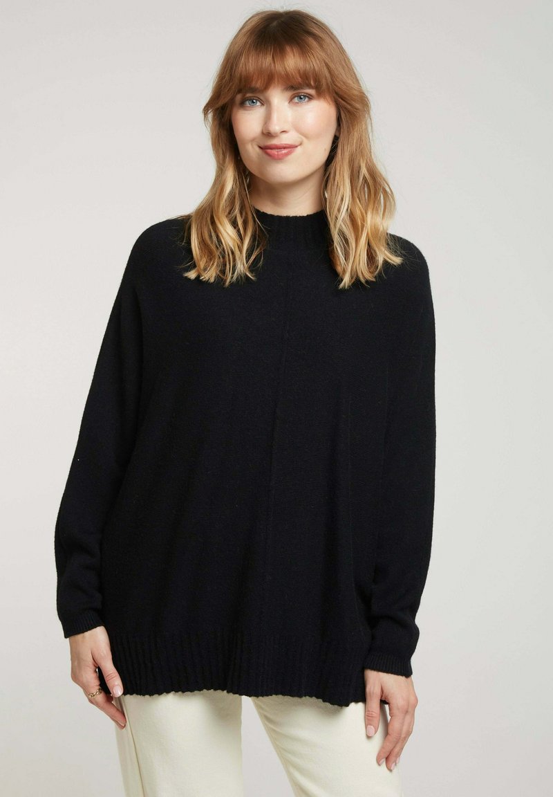 Pull noir oversize avec un col montant et des poignets côtelés. Texture douce et design à épaules tombantes, associé à un pantalon de couleur claire.