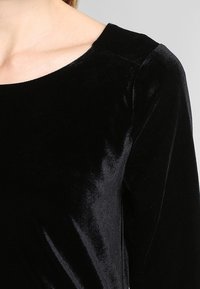 Haut en velours noir à la texture lisse, col rond et manches longues. Il présente une coupe ajustée et une brillance subtile.