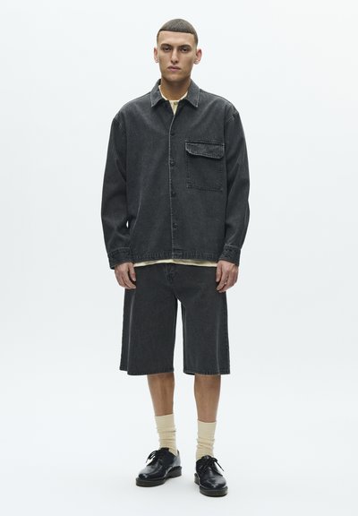 Tamno traper jakna s prednjim džepom, uparena s odgovarajućim tamnim traper kratkim hlačama. Model nosi bež čarape i crne cipele, postavljen na jednobojnu pozadinu.