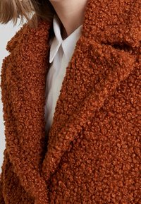Texturerad orange kappa med ett fluffigt, boucle-tyg; har en bred slag och närbild av en vit skjorta med krage under.