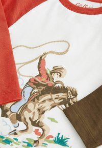 Witte T-shirt met rode en olijfgroene raglanmouwen; bevat een cowboy op een bokkende hengst, bedrukt in bruine en groene tinten met touwdetail.