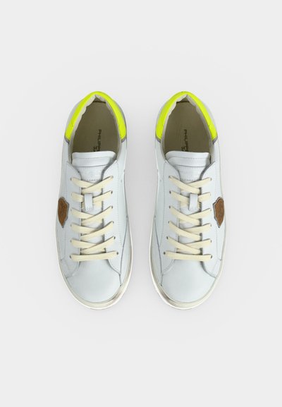 Paire de baskets blanches à lacets avec des lacets beiges, des patchs logo marron sur les côtés et un détail jaune fluo au niveau des talons, vues de dessus.