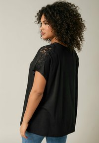 Maglietta nera a maniche corte con dettagli in pizzo intricati sulle spalle, vestibilità rilassata e tessuto morbido. Indossata con jeans blu.