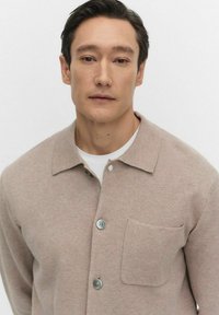 Beige gebreide shirt met een kraag, voorzien van een knoopsluiting aan de voorkant, een enkel borstzakje en lichtblauwe knopen tegen een effen achtergrond.