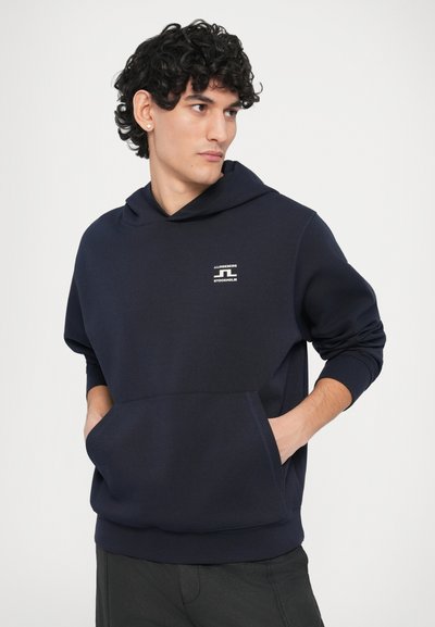 Μπλε ναυτικό φούτερ με μπροστινή τσέπη, κουκούλα με ρυθμιζόμενο κορδόνι και μικρό λογότυπο στο στήθος. Το ύφασμα φαίνεται μαλακό και λείο.