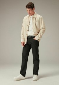 Beige corduroy shirt over een wit t-shirt, zwarte pants en witte sneakers. Het shirt heeft een zak en knoopsluiting, en heeft een textuur op het oppervlak.