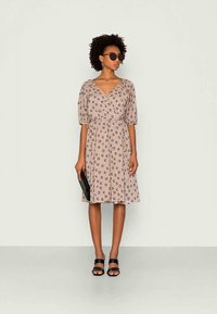 Vestido bege com padrão floral, decote em V, mangas bufantes e cintura ajustada, combinado com acessórios e sandálias pretas.