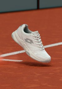 Λευκό αθλητικό παπούτσι τένις από συνθετικό υλικό, με υφασμένη επιφάνεια, στρογγυλή μύτη και ανοιχτό γκρι λεπτομέρειες, πάνω σε γήπεδο με κόκκινο χώμα.