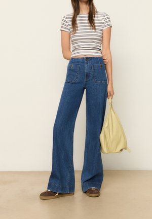 High-waisted blue flared jeans with two front pockets, paired with a striped crop top and a yellow bag. Brown shoes complete the outfit.