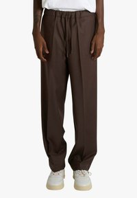 Pantaloni marroni a gamba larga con vita elastica e coulisse, dalla texture liscia, abbinati a sneakers bianche.