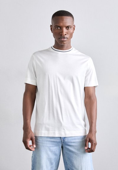 T-shirt bianco a maniche corte con colletto a costine bianco e nero; tessuto di cotone; vestibilità comoda. Indossato con pantaloni corti di jeans azzurri.