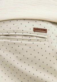 Poche de pantalon beige à motifs avec une petite étiquette en cuir marron marquée "JACK&JONES" et un tissu blanc texturé au-dessus.