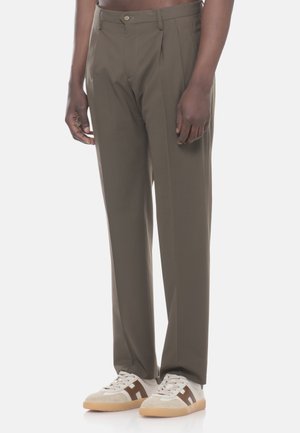Persona che indossa pantaloni eleganti verde oliva a taglio ampio e sneakers bianche con dettagli marroni, in piedi su uno sfondo chiaro e uniforme.