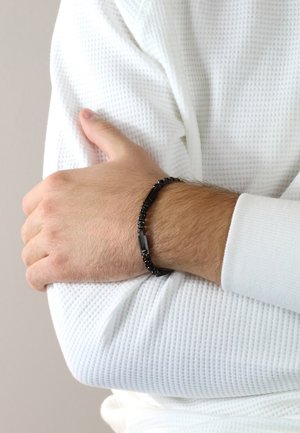 Bracelet made of black beads with rectangular accents, worn on a light-skinned arm, with a white textured long-sleeve shirt visible.