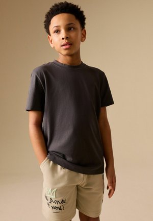 Junge steht mit den Händen in den Taschen, trägt ein dunkelgraues T-Shirt und beige Shorts mit einer mintfarbenen Krone und schwarzem Graffiti-Text.