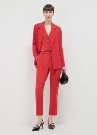 Roter maßgeschneiderter Anzug mit einem taillierten Blazer und einer Weste mit Knöpfen, kombiniert mit schmal geschnittenen Hosen und schwarzen High Heels. Kleine schwarze Handtasche enthalten.