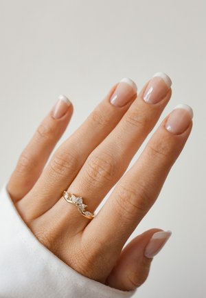 Anello d'oro a forma di onda con più pietre chiare, posato su una mano con unghie naturali smaltate in nude con punte francesi.