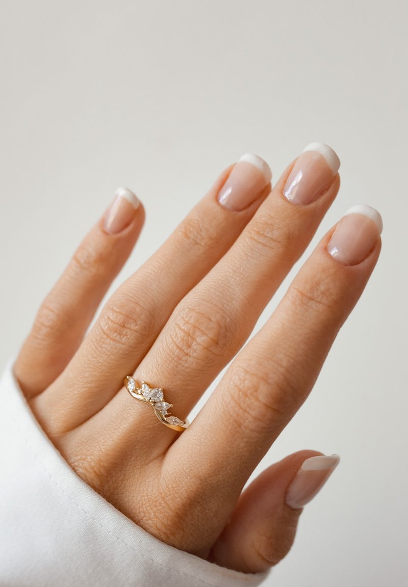Złota pierścionek falowy z wieloma przezroczystymi kamieniami, umieszczony na dłoni z naturalnymi paznokciami pokrytymi nudowym lakierem i francuskimi końcówkami.