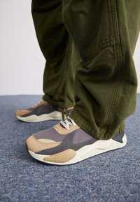 Zapatillas deportivas con una parte superior de malla gris, acentos de cuero color marrón y una suela texturizada, combinadas con pantalones holgados de color verde olivo sobre una moqueta azul.