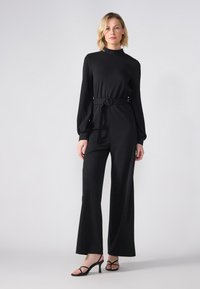 Black jumpsuit with a high neckline, long sleeves, and a belted waist. Flared pants and smooth fabric, paired with strappy black heels.