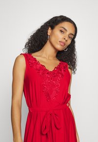 Vestido vermelho com padrões florais bordados na gola em V, cintura ajustada com um cinto atado, tecido suave e design sem mangas.