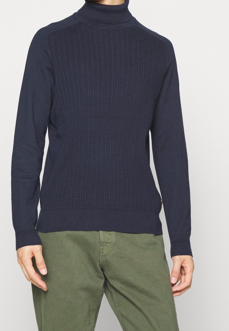 Navyblå polotröja med ribbad textur på framstycket, kontrasterande släta ärmar och en figurnära design. I kombination med olivgröna byxor.