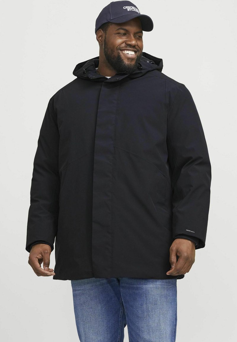 Chaqueta impermeable negra con capucha, con cierre de cremallera en el frente, bolsillos y un ajuste relajado. Lleva puesta sobre unos vaqueros azules, combinada con una gorra azul marino.