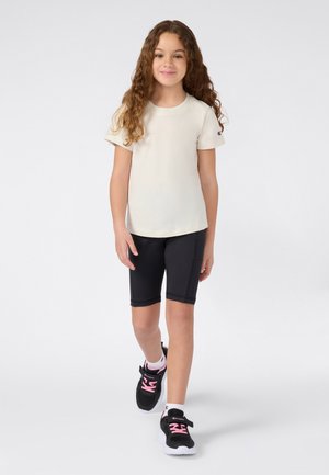 Jeune fille aux cheveux bouclés portant un t-shirt blanc, un short de vélo noir et des baskets noires avec des lacets roses marchant vers l'avant sur un fond uni.