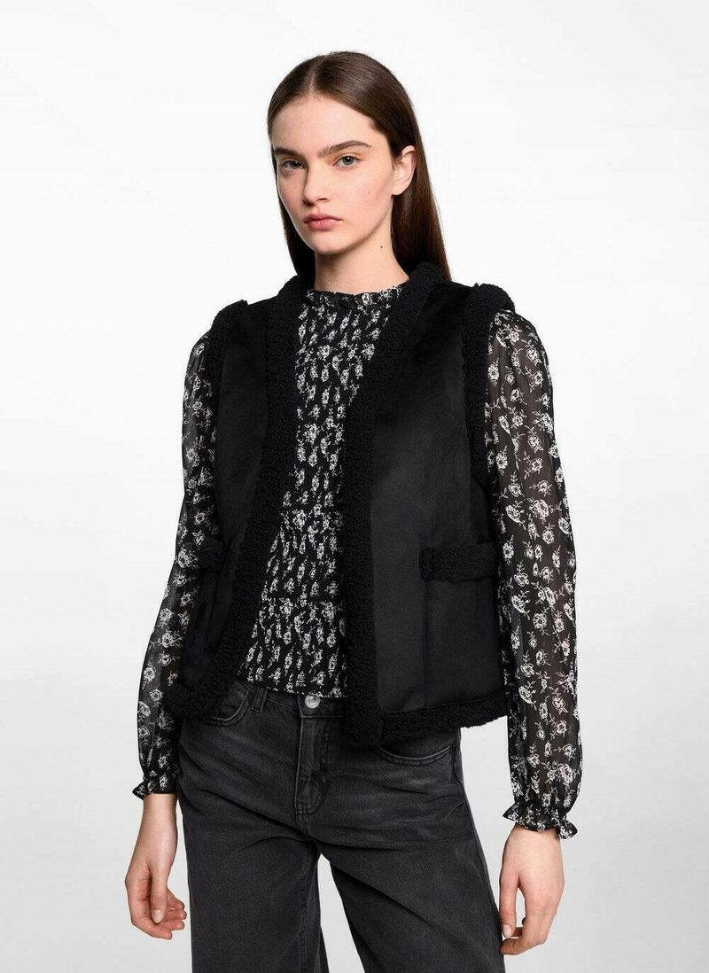 Svart väst med texturerad trim, med rundad halsringning, utan knäppningar och två framfickor, lager på lager över en mönstrad blus.