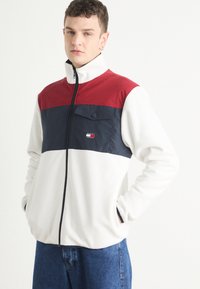 Giacca in pile con colletto alto, caratterizzata da una parte superiore rossa, una parte centrale blu navy e una parte inferiore bianca. Include una tasca sul petto e chiusura con zip.