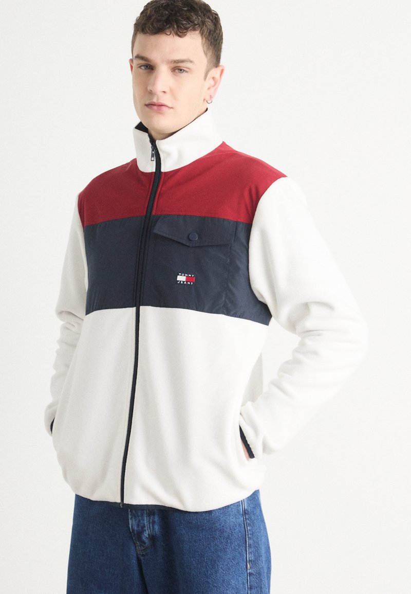 Giacca in pile con colletto alto, caratterizzata da una parte superiore rossa, una parte centrale blu navy e una parte inferiore bianca. Include una tasca sul petto e chiusura con zip.