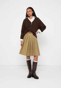 Žena oblečená v hnedom pletenom svetri, bielej košeli s golierom, béžovej plisovanej sukni, hnedom opasku a hnedých čižmách po kolená s bielymi ponožkami.