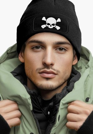 Black knit beanie with a white skull and crossbones patch, worn with a green puffer jacket. Soft texture, close-fitting design.