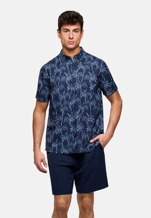 Hombre vestido con camisa de manga corta de botones azul marino con estampado de palmeras en azul claro y pantalones cortos azul marino sólidos, de pie con la mano en el bolsillo.