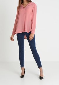 Blouse rose à manches longues avec col et encolure en V, associée à un jean skinny bleu foncé et des chaussures à talons hauts noires.