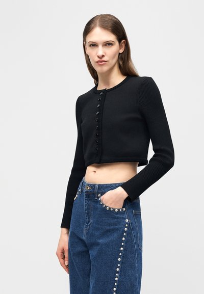 Mujer que lleva un suéter negro abotonado y corto y unos vaqueros de cintura alta azules con tachuelas tipo perla a lo largo de los bolsillos y las costuras exteriores.
