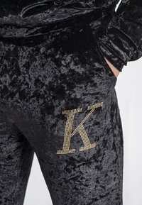 Zwarte fluwelen joggers met een opvallende goud-gestudeerde letter 'K' op de linkerzak. De stof heeft een zachte, gestructureerde uitstraling.