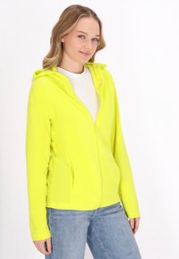Felpa zip-up gialla brillante con cappuccio, dotata di tasche laterali e un tessuto leggero e morbido. Indossata sopra una maglietta bianca e jeans.