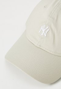 Beige Basecap mit weißem gesticktem, ineinandergreifendem "NY"-Logo auf der Vorderseite, vor einem hellen, einfarbigen Hintergrund dargestellt.