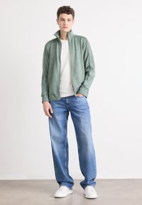 Lysegrønn zip-up jakke med høy krage, kombinert med en tettsittende hvit t-skjorte og løse blå jeans. Hvite sneakers fullfører antrekket.