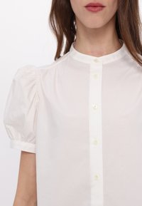 Femme portant un chemisier blanc avec des manches courtes bouffantes et un col mandarin boutonné, lèvres visibles mais pas d'autres traits du visage.