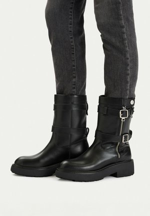 Botas negras de cuero hasta la mitad de la pantorrilla con suelas gruesas, cremallera lateral y correas con hebillas, usadas con jeans ajustados de color gris oscuro.