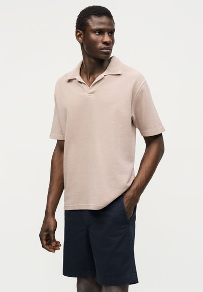Homme portant un polo beige à manches courtes et un short bleu marine, debout avec une main dans la poche, regardant de côté.