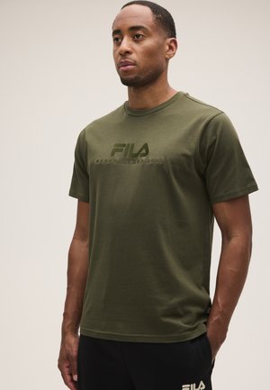 Mann trägt ein olivgrünes FILA-T-Shirt und schwarze Hose, steht vor einem schlichten hellen Hintergrund mit den Händen in den Taschen.