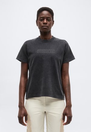 Person med kort hår iført en falmet sort "66 North Iceland" t-shirt og lyse bukser, stående mod en ensfarvet baggrund.