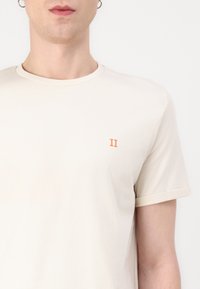 Tricou cremos cu mânecă scurtă, confecționat din bumbac moale, având brodat pe partea stângă a pieptului un mic "II" portocaliu.