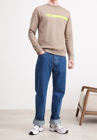 Beige sweatshirt med ett horisontellt neon-gult streck och texten "BOSS", kombinerad med blå denimjeans. Modellen bär vita sneakers.